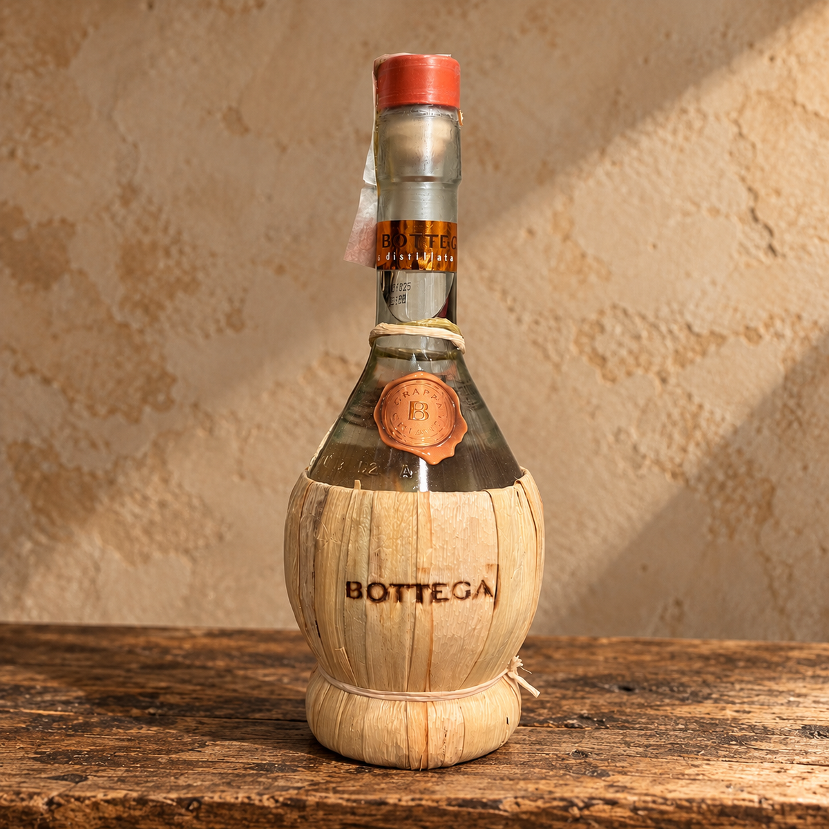 Grappa Bottega italiana in bottiglia di vetro rivestita in paglia naturale, su tavolo rustico in legno – Essenza Pisterzi.